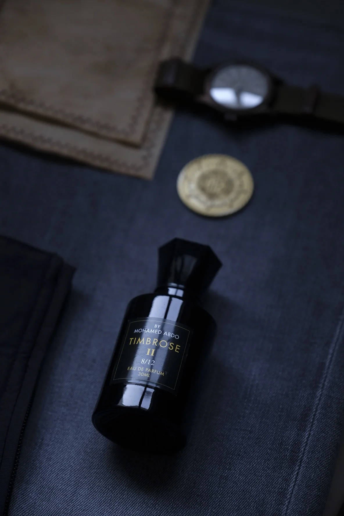 Timbrose II lying on dark blue denim beside a leather wallet, a wristwatch and a gold coin