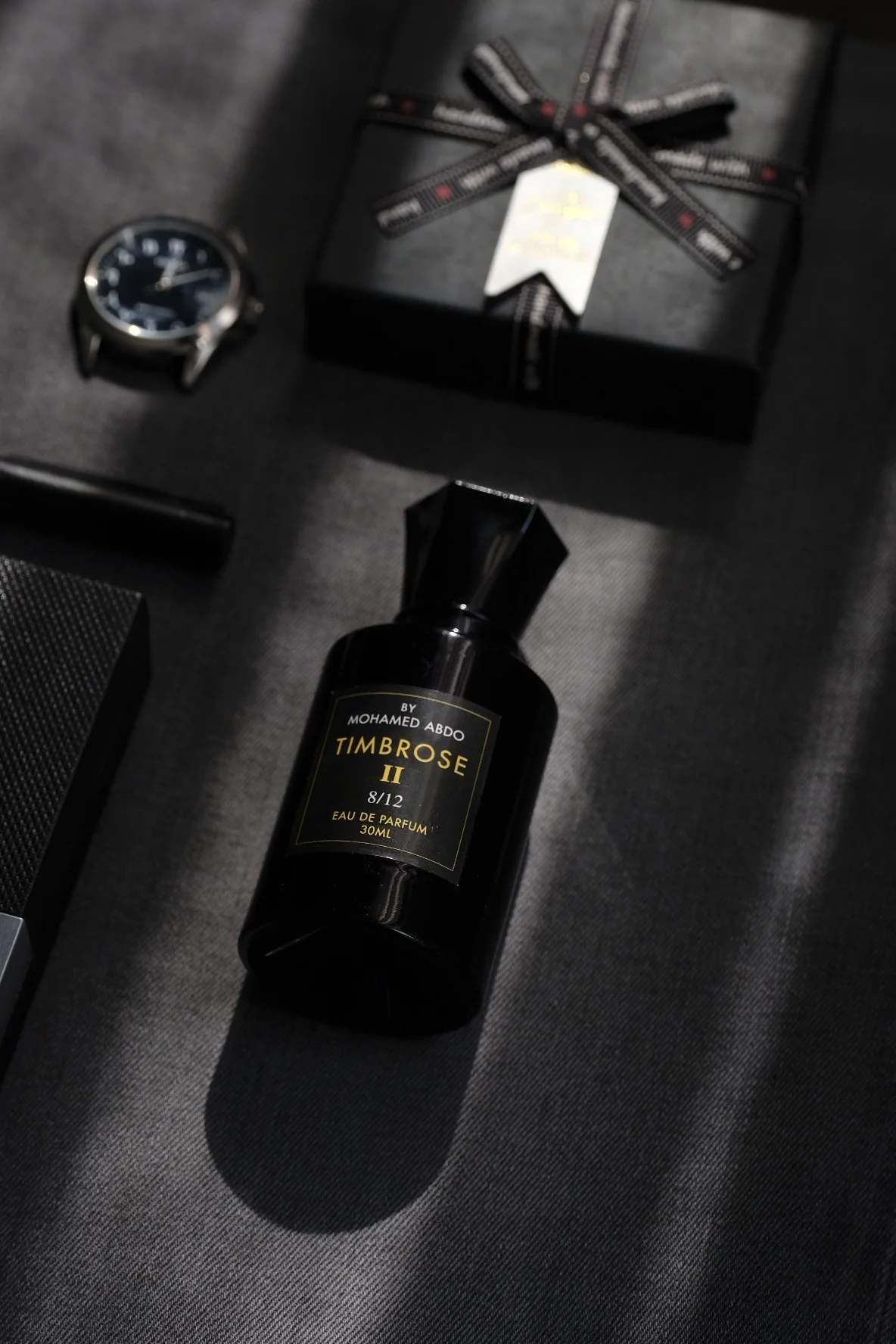 Timbrose II in a slant of sunlight on grey cloth beside a wristwatch and a black gift box tied with ribbon