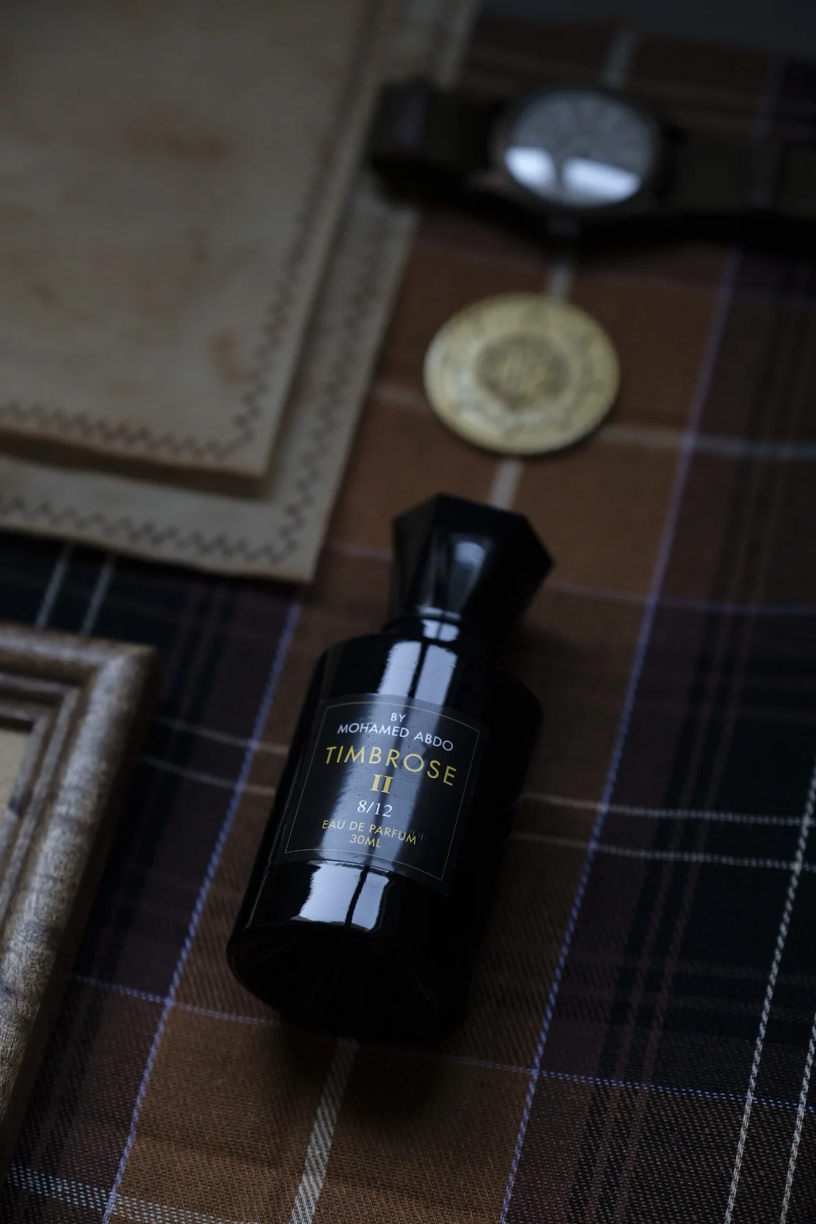 Timbrose II lying on brown tartan cloth beside a leather wallet, a wristwatch, a gold coin and a wooden frame